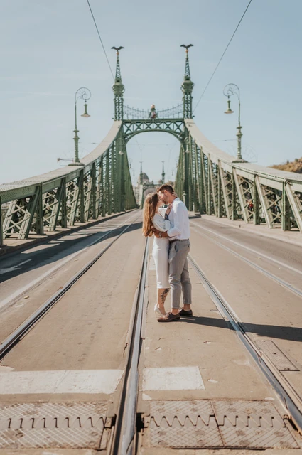 The couple and Liberty Bridge’s long perspective | Budapest engagement photoshoot A wide portrait of the couple together with the green towers and long perspective of Liberty Bridge behind them during their engagement photoshoot in Budapest.