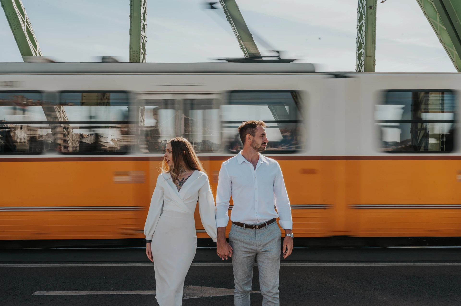 A yellow tram behind their bridge portraits | Budapest engagement photoshoot A wide view of the couple holding hands on Liberty Bridge while a yellow tram passes behind them during their engagement photoshoot in Budapest.