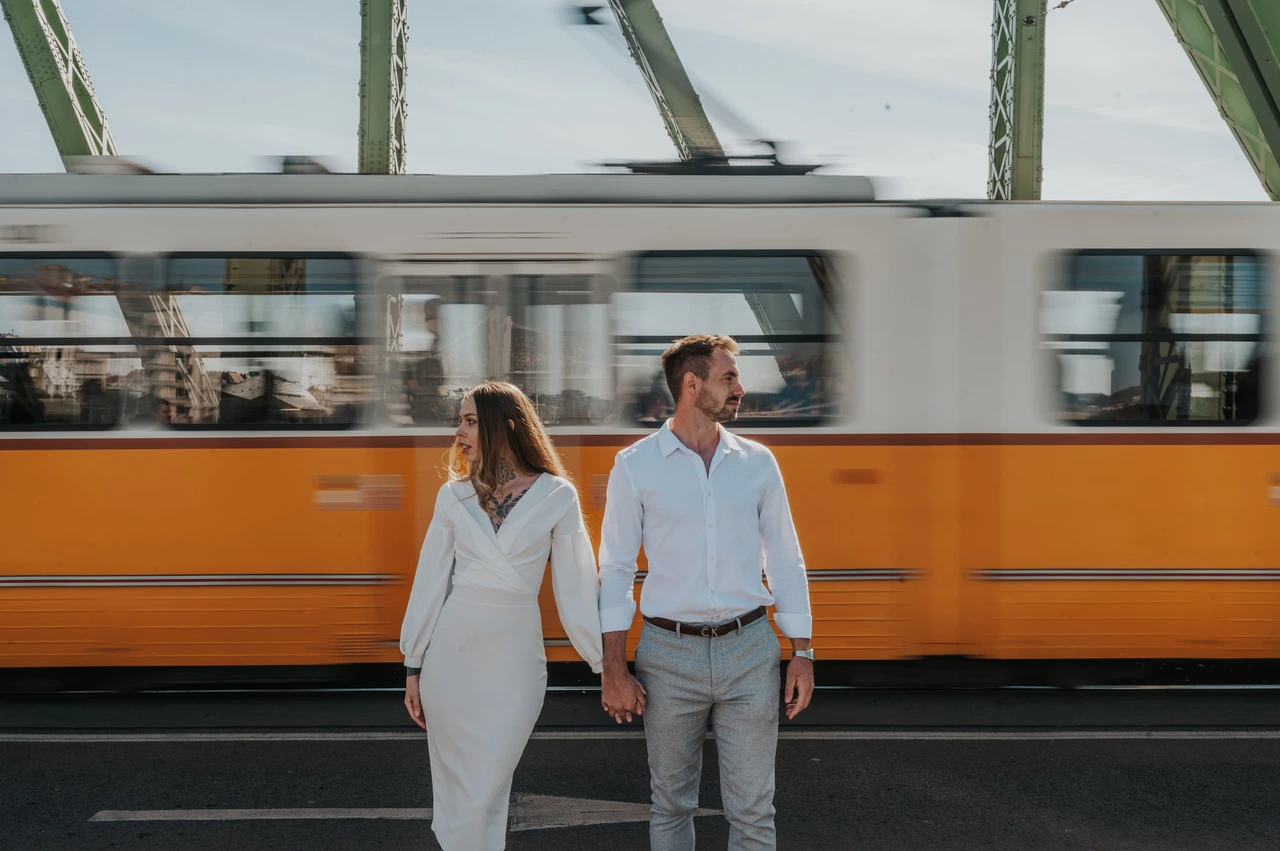 A yellow tram behind their bridge portraits | Budapest engagement photoshoot A wide view of the couple holding hands on Liberty Bridge while a yellow tram passes behind them during their engagement photoshoot in Budapest.