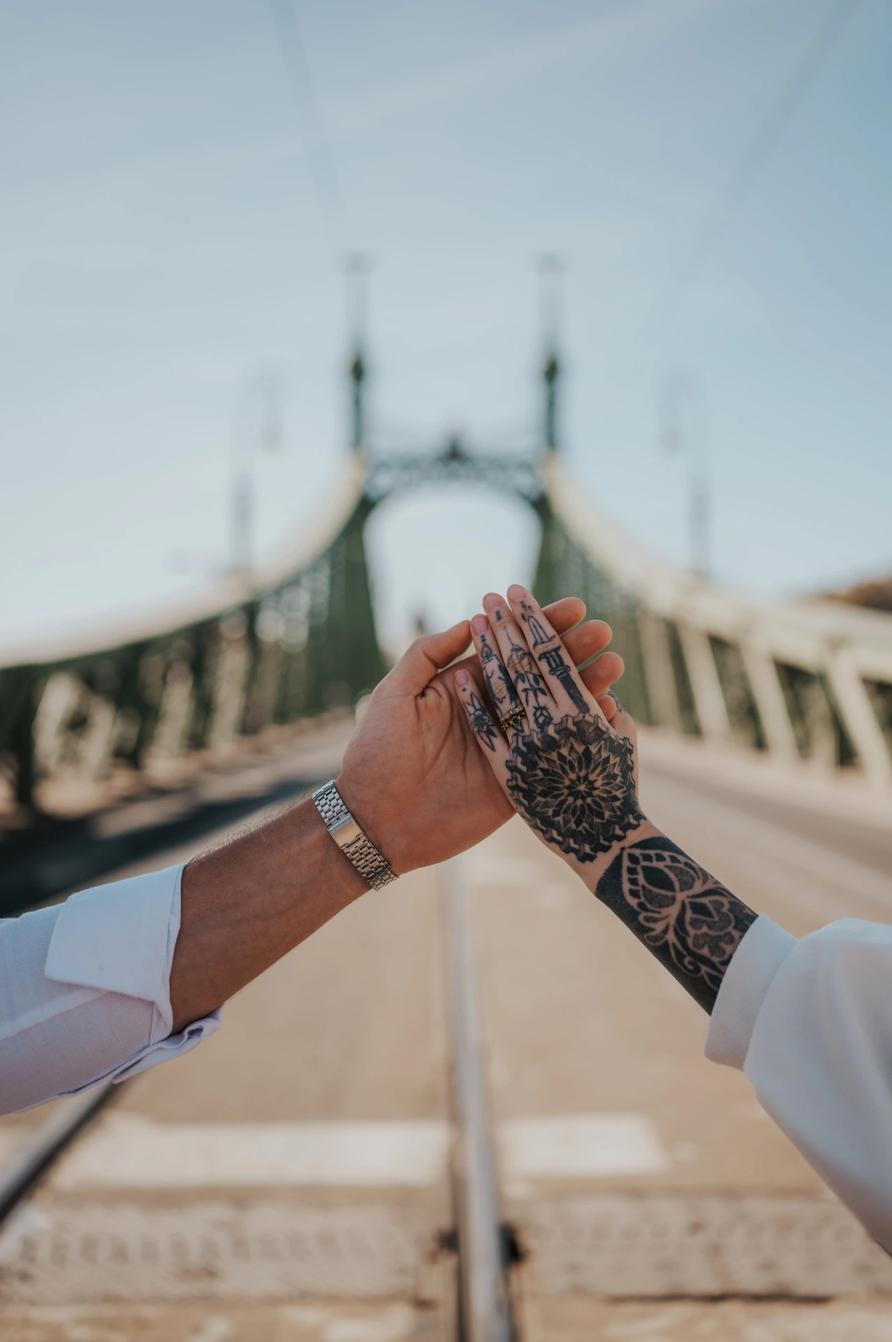 A close-up of the couple’s hands together showing an engagement ring and tattoos, with Liberty Bridge softly blurred behind them during their engagement photoshoot in Budapest.