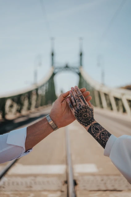 A close-up of the couple’s hands together showing an engagement ring and tattoos, with Liberty Bridge softly blurred behind them during their engagement photoshoot in Budapest.