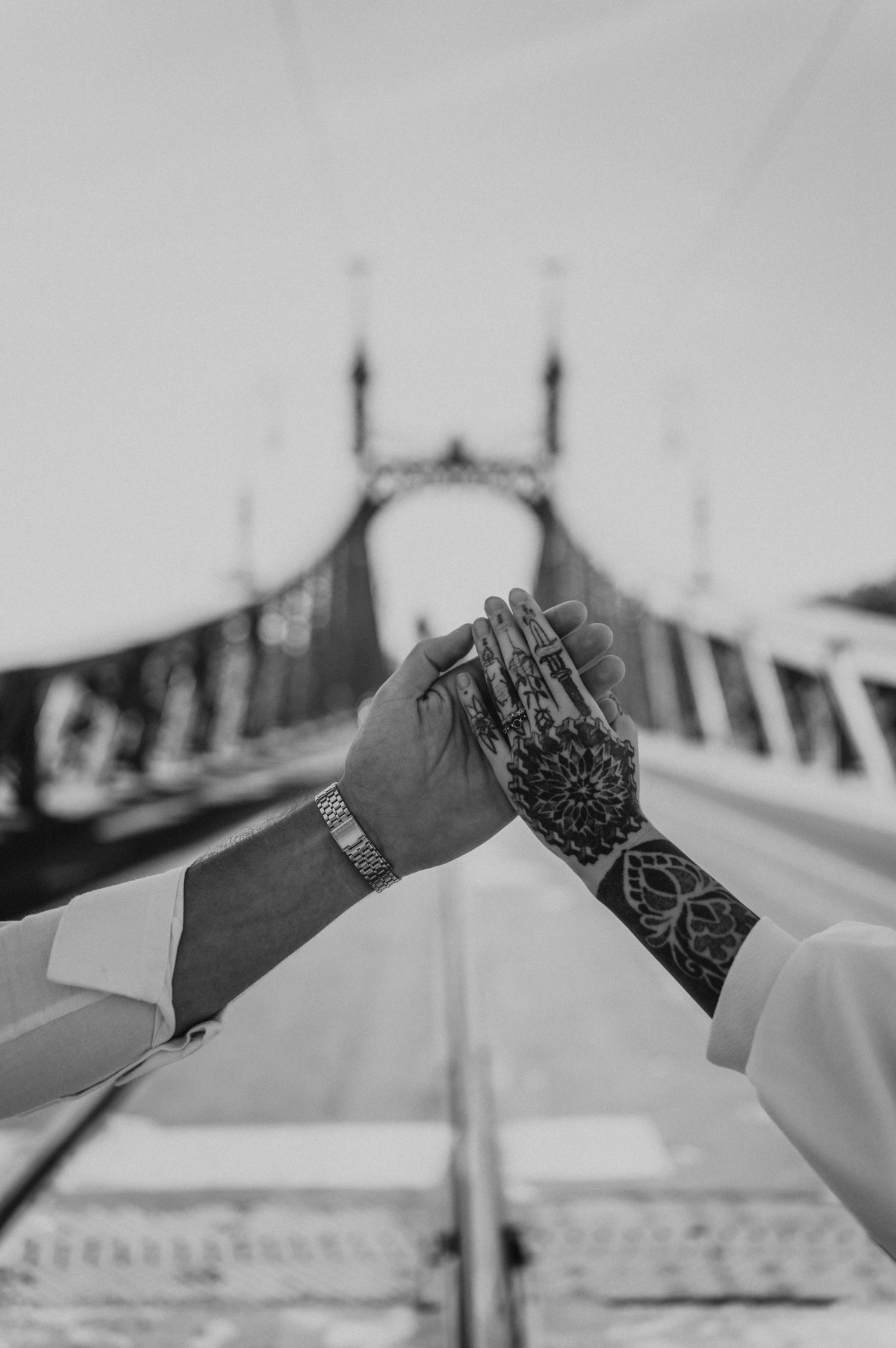 Black-and-white photograph of a close-up of the couple’s hands showing their engagement ring and tattoos, with Liberty Bridge softly blurred behind them during their engagement photoshoot in Budapest.