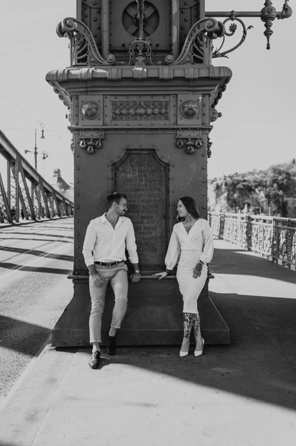 The ornate bridge details in black and white | Budapest engagement photoshoot Black-and-white photograph of the couple standing together beside an ornate lamp-post base on Liberty Bridge during their engagement photoshoot in Budapest.