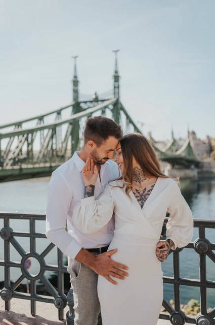 The engaged couple embracing beside the Danube with Liberty Bridge behind them during their engagement photoshoot in Budapest.