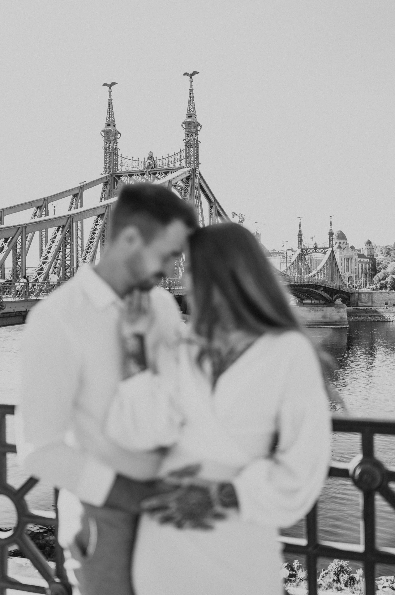 Black-and-white photograph of a softly focused portrait of the couple leaning together beside the riverside railing with Liberty Bridge behind them during their engagement photoshoot in Budapest.