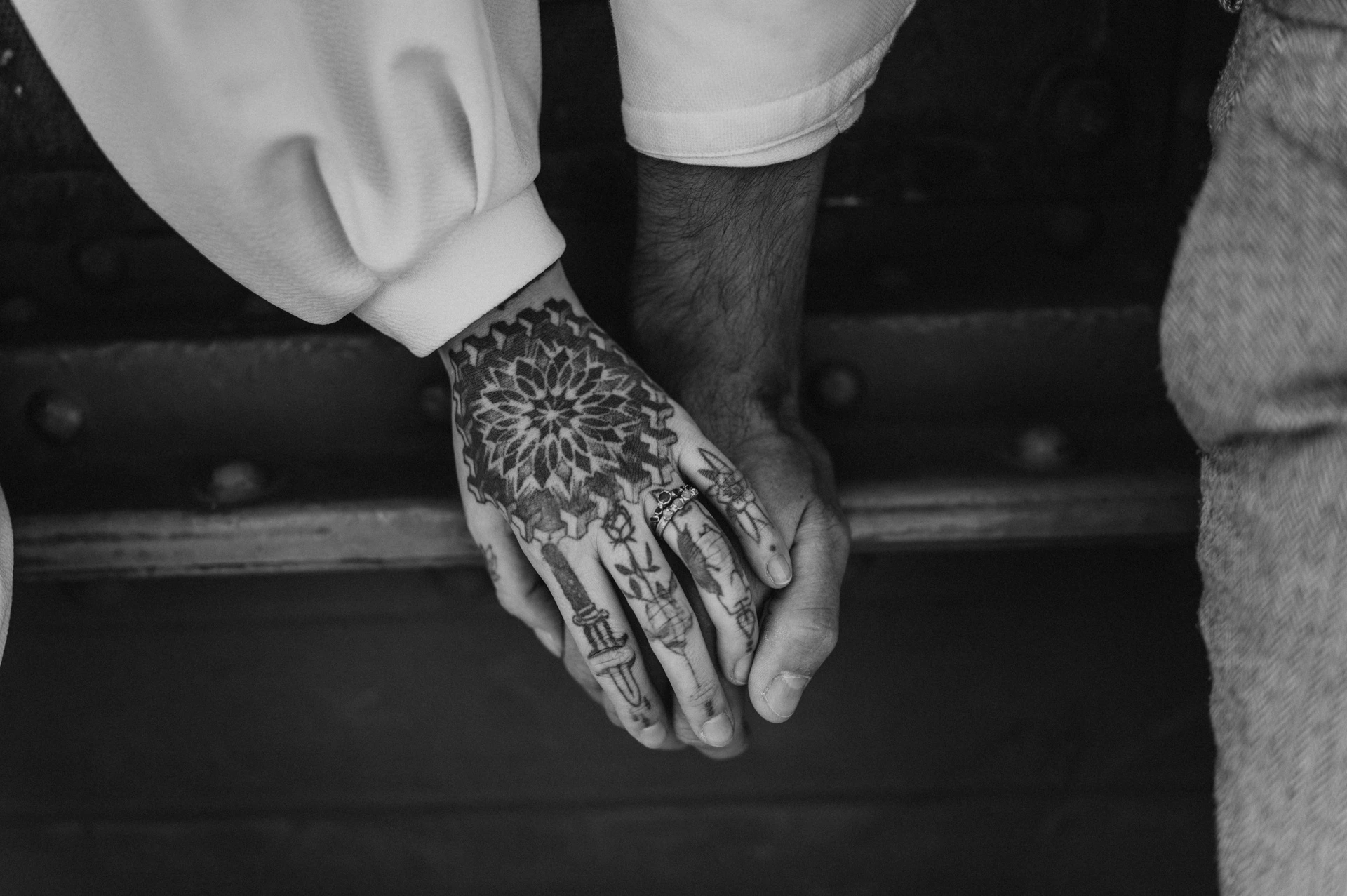 Black-and-white photograph of a wide close-up of the couple’s tattooed hands resting together against the bridge steelwork during their engagement photoshoot in Budapest.