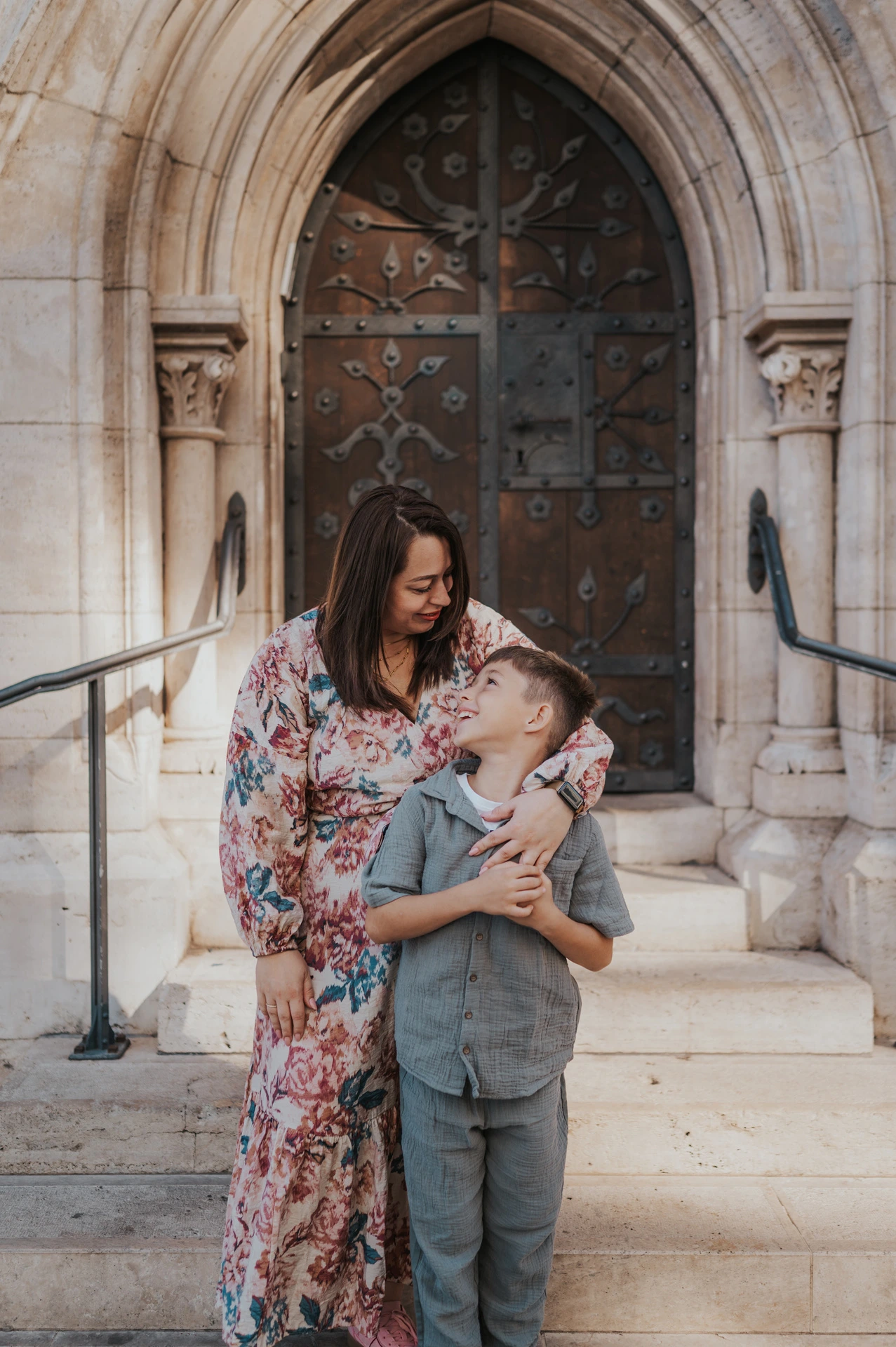 A mother kissing her son’s head as they stand together on the steps of a carved stone doorway during their family photoshoot in Budapest.