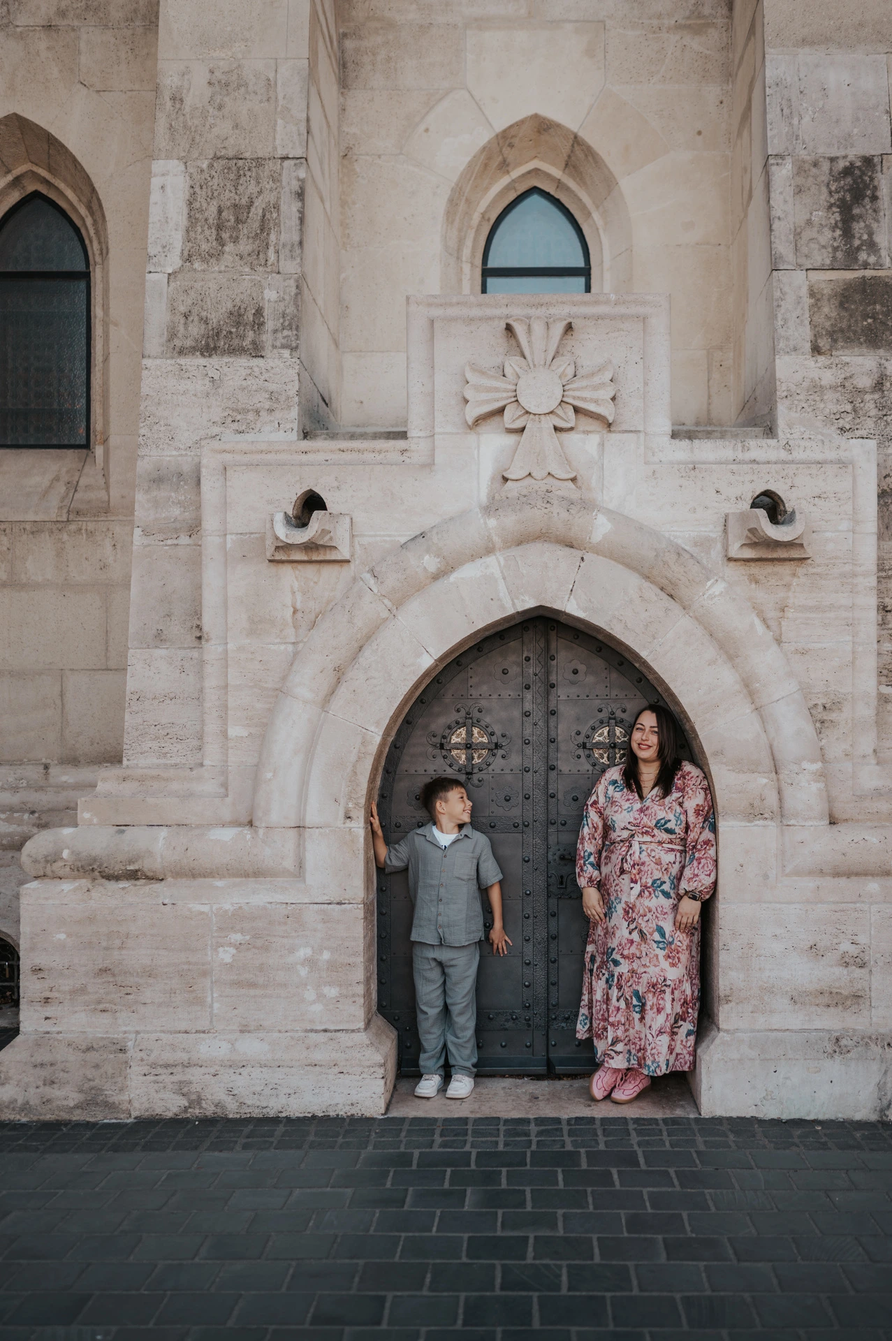 A mother in a floral dress looking towards her son as they stand beside a small arched stone doorway during their family photoshoot in Budapest.