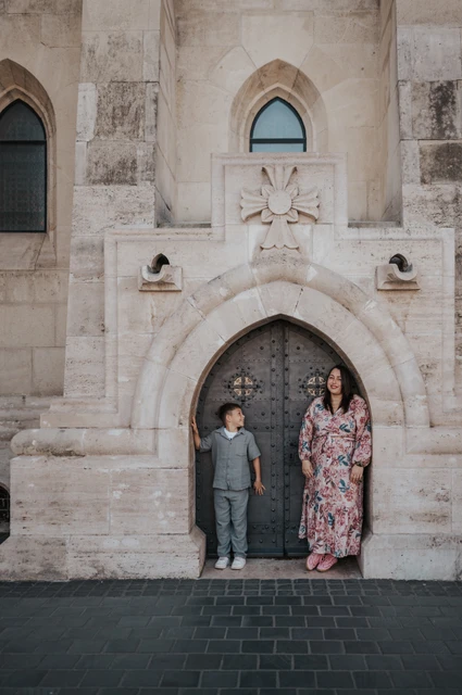 Together beside the little arched door | Budapest family photoshoot A mother in a floral dress looking towards her son as they stand beside a small arched stone doorway during their family photoshoot in Budapest.