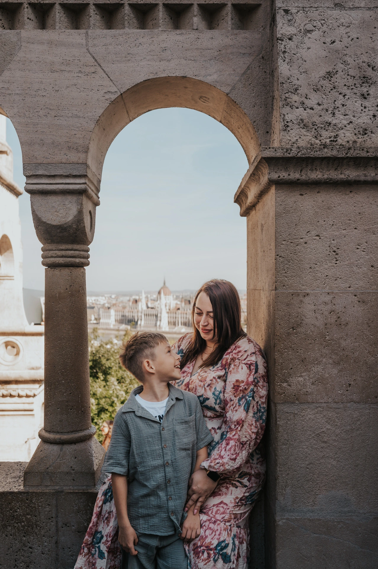 A mother and son smiling at one another beneath a stone arch at Fisherman’s Bastion, with Parliament visible across the river during their family photoshoot in Budapest.