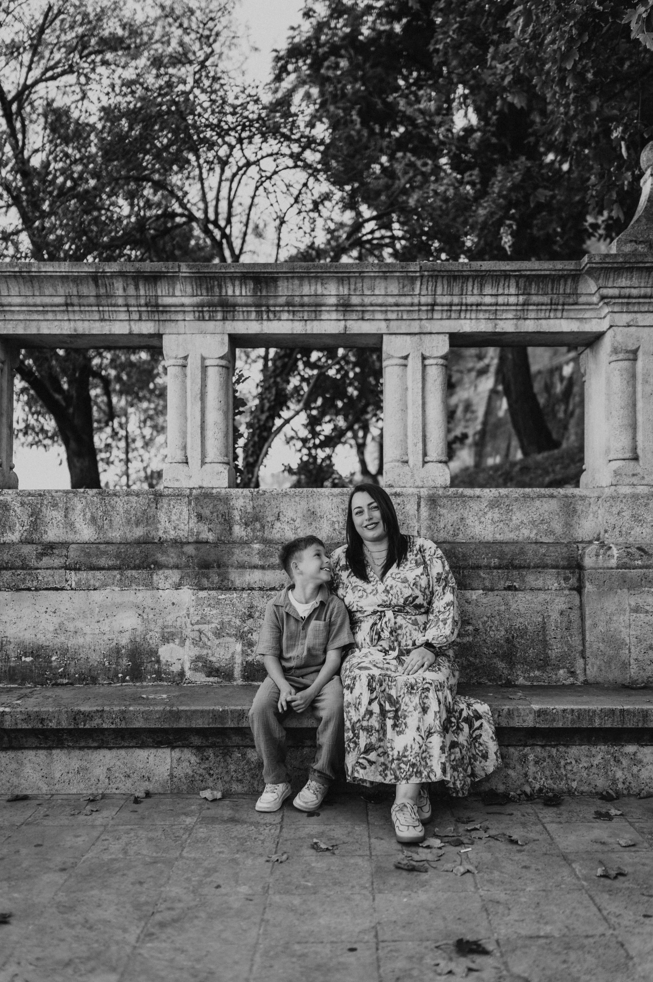 Black-and-white photograph of a mother and son sitting close together on a stone bench in front of a carved balustrade and leafy trees during their family photoshoot in Budapest.