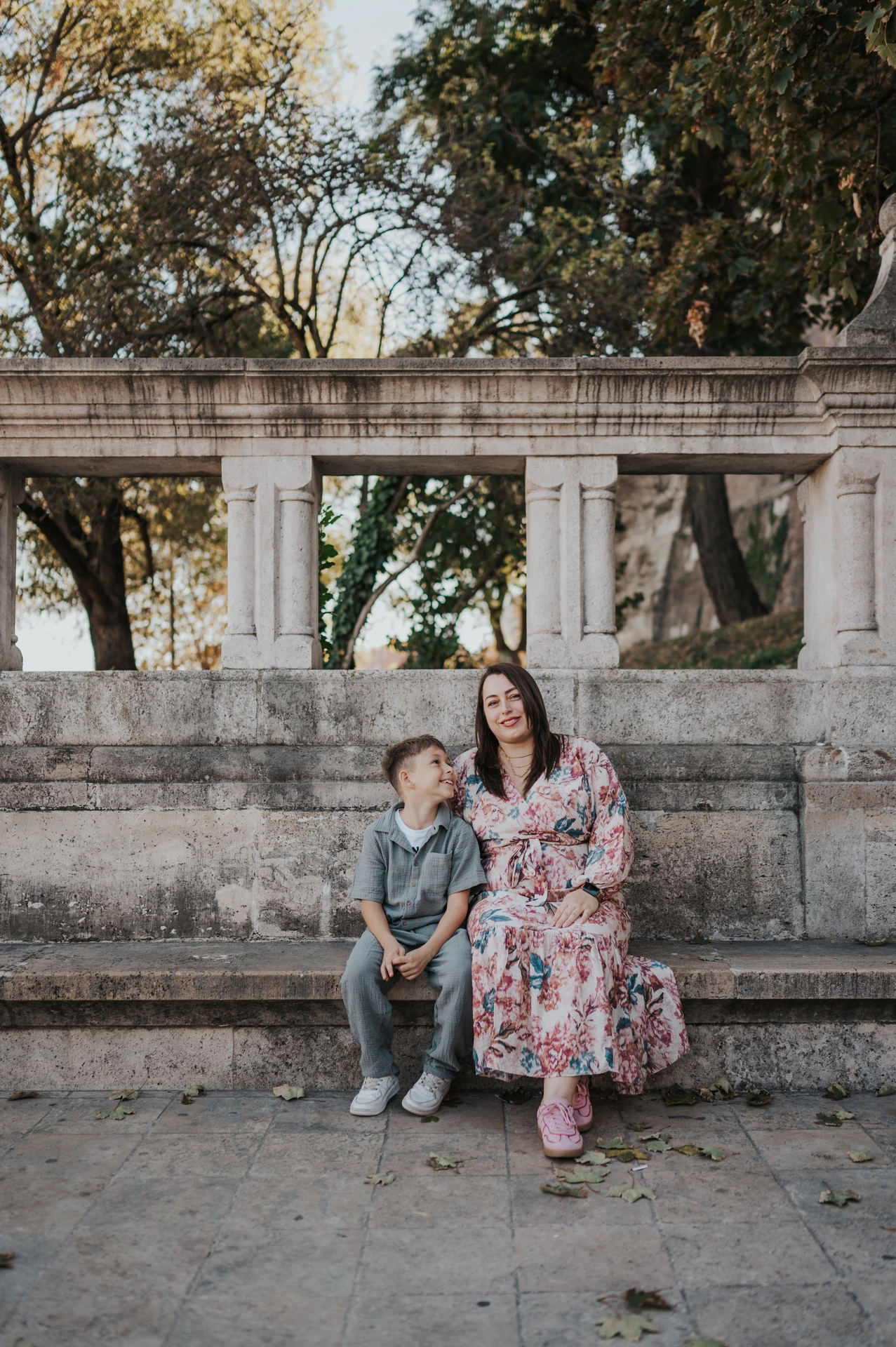 A mother and son smiling at one another as they sit together on a stone bench beneath leafy trees during their family photoshoot in Budapest.