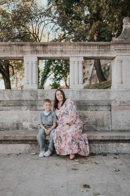 A mother and son sitting together on a stone bench, with a carved balustrade and leafy trees behind them during their family photoshoot in Budapest.