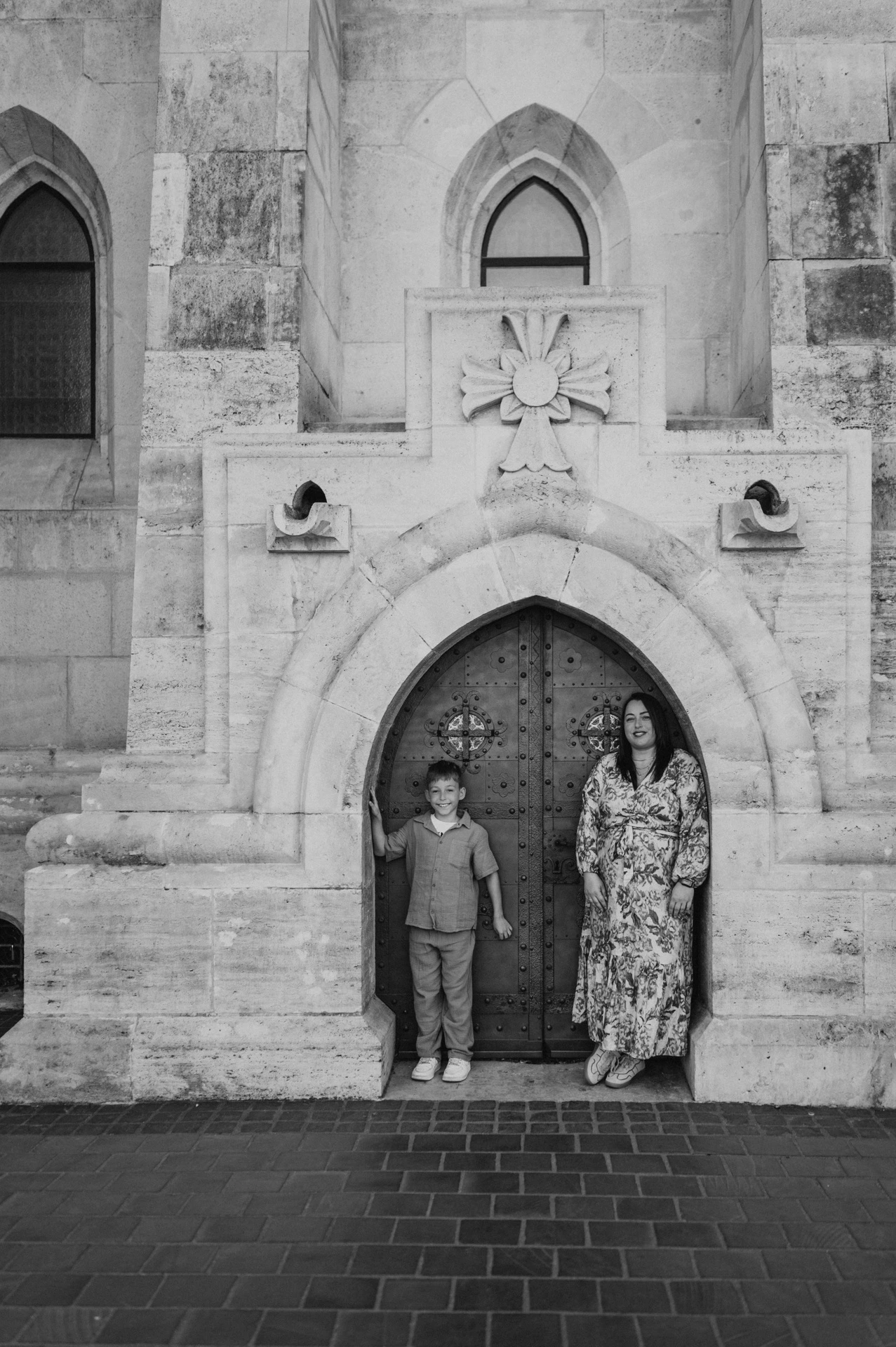 Black-and-white photograph of a mother and son standing together in front of a small arched doorway set into a pale stone wall during their family photoshoot in Budapest.