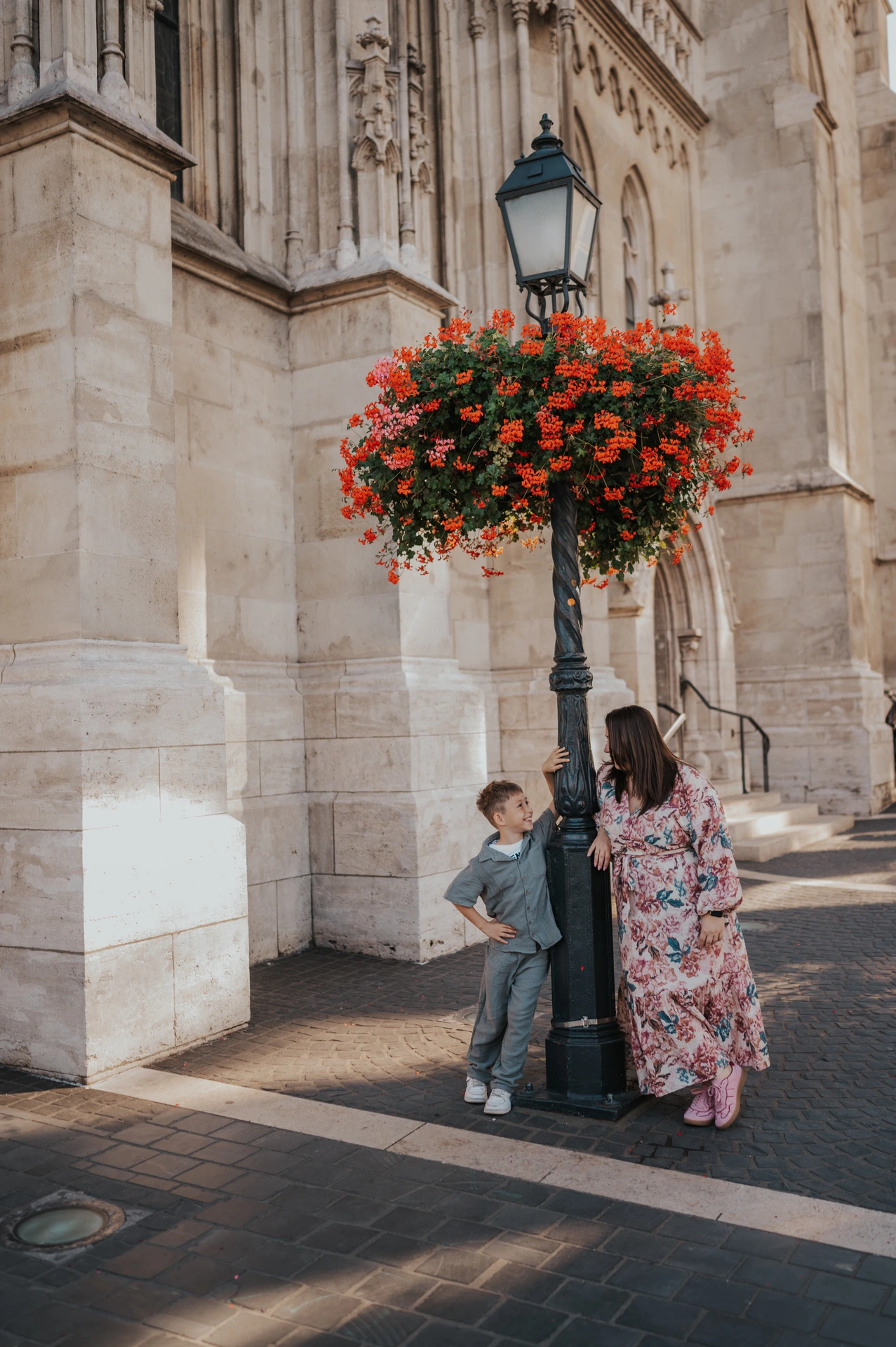A mother leaning towards her smiling son beside a street lamp and a hanging basket of red flowers during their family photoshoot in Budapest.