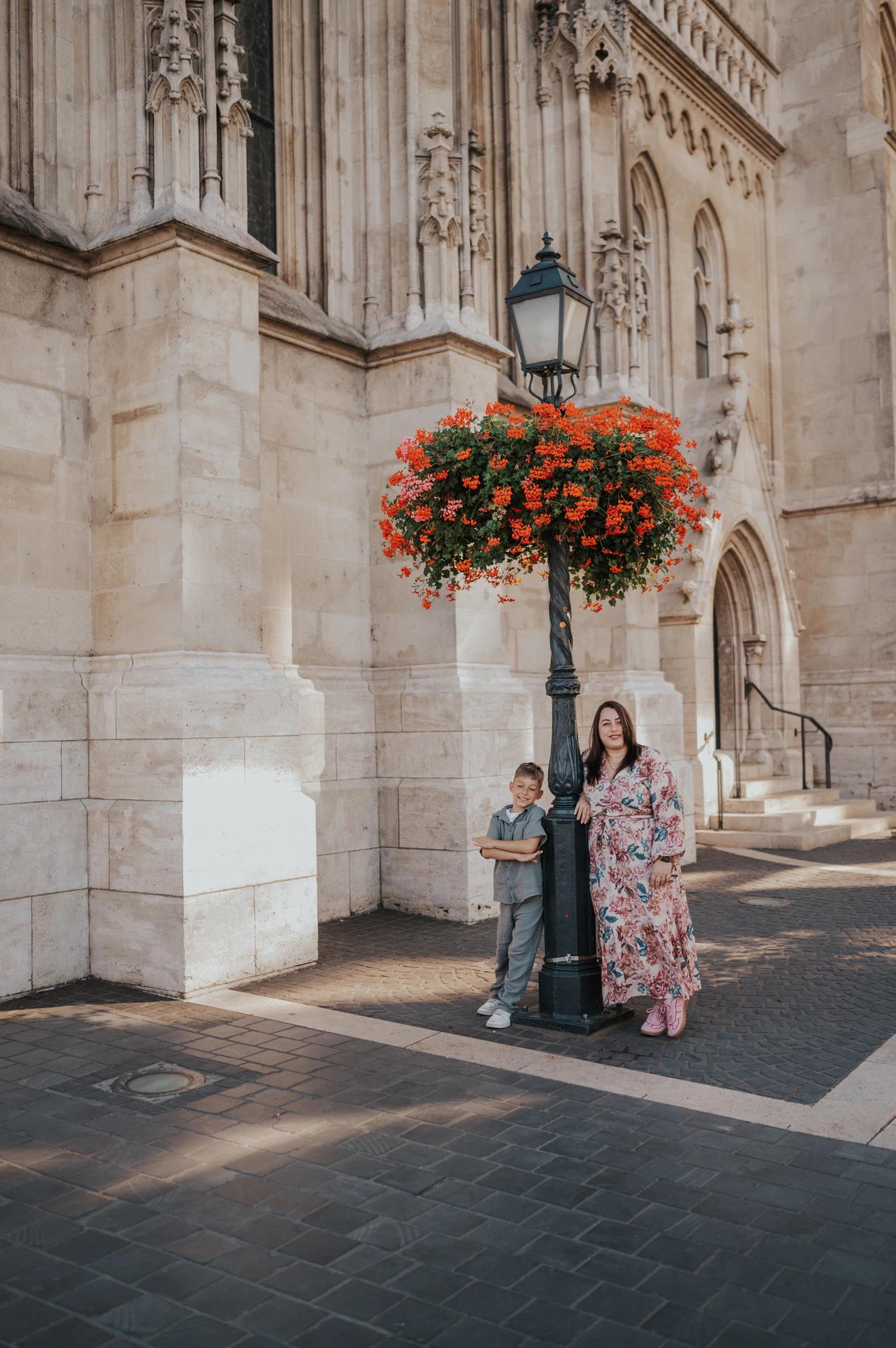 A mother and son standing together beside a street lamp and a hanging basket of red flowers against a pale stone building during their family photoshoot in Budapest.