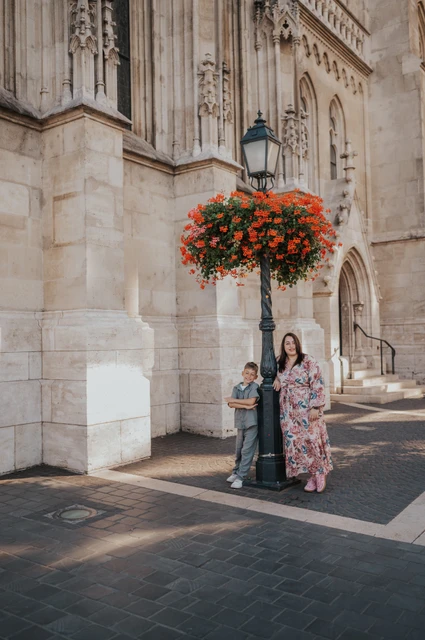 A mother and son standing together beside a street lamp and a hanging basket of red flowers against a pale stone building during their family photoshoot in Budapest.