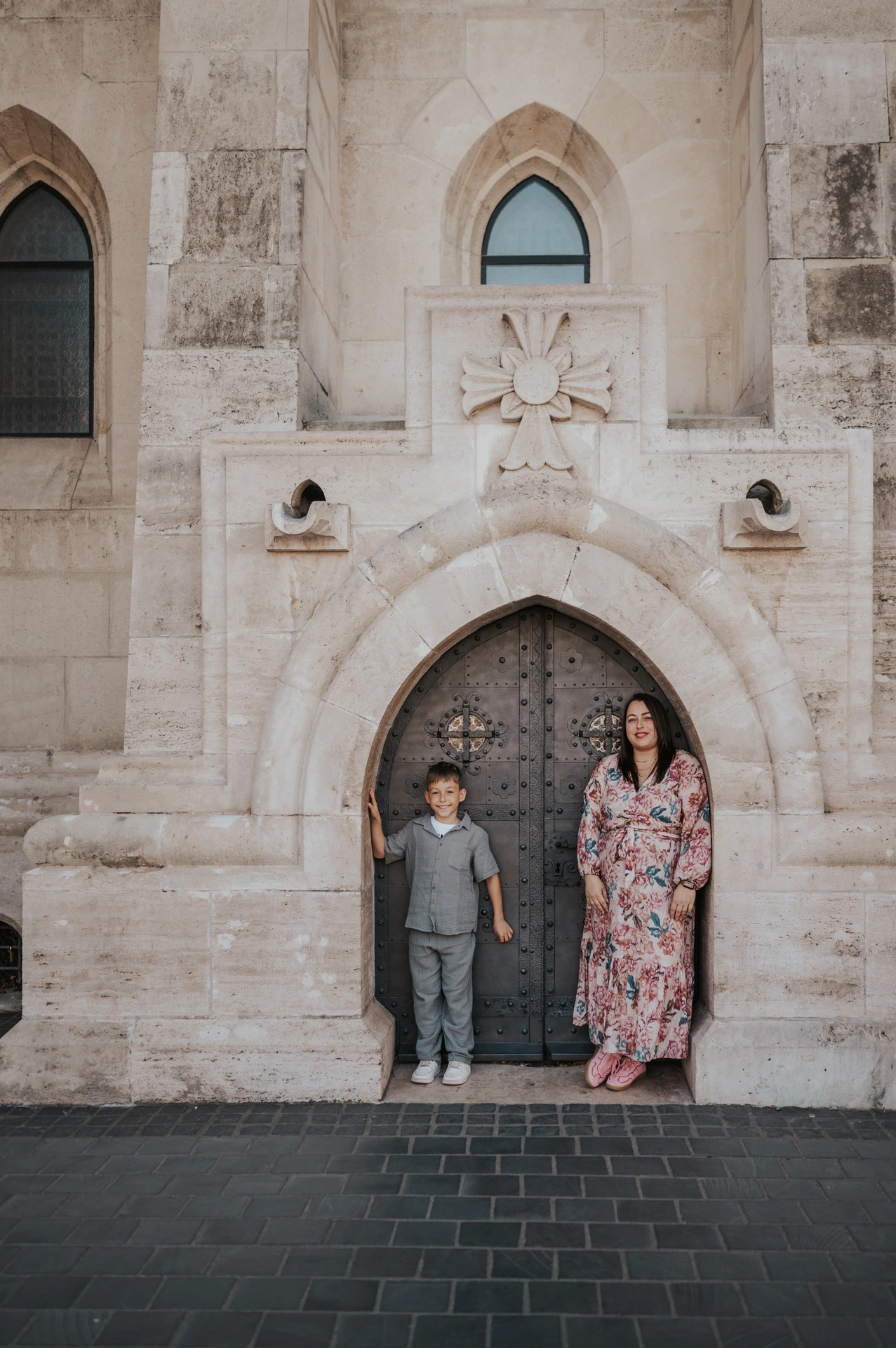 A mother and son standing side by side in front of a small arched doorway set into a pale stone wall during their family photoshoot in Budapest.