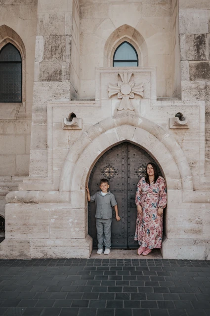 Side by side at a little stone doorway | Budapest family photoshoot A mother and son standing side by side in front of a small arched doorway set into a pale stone wall during their family photoshoot in Budapest.