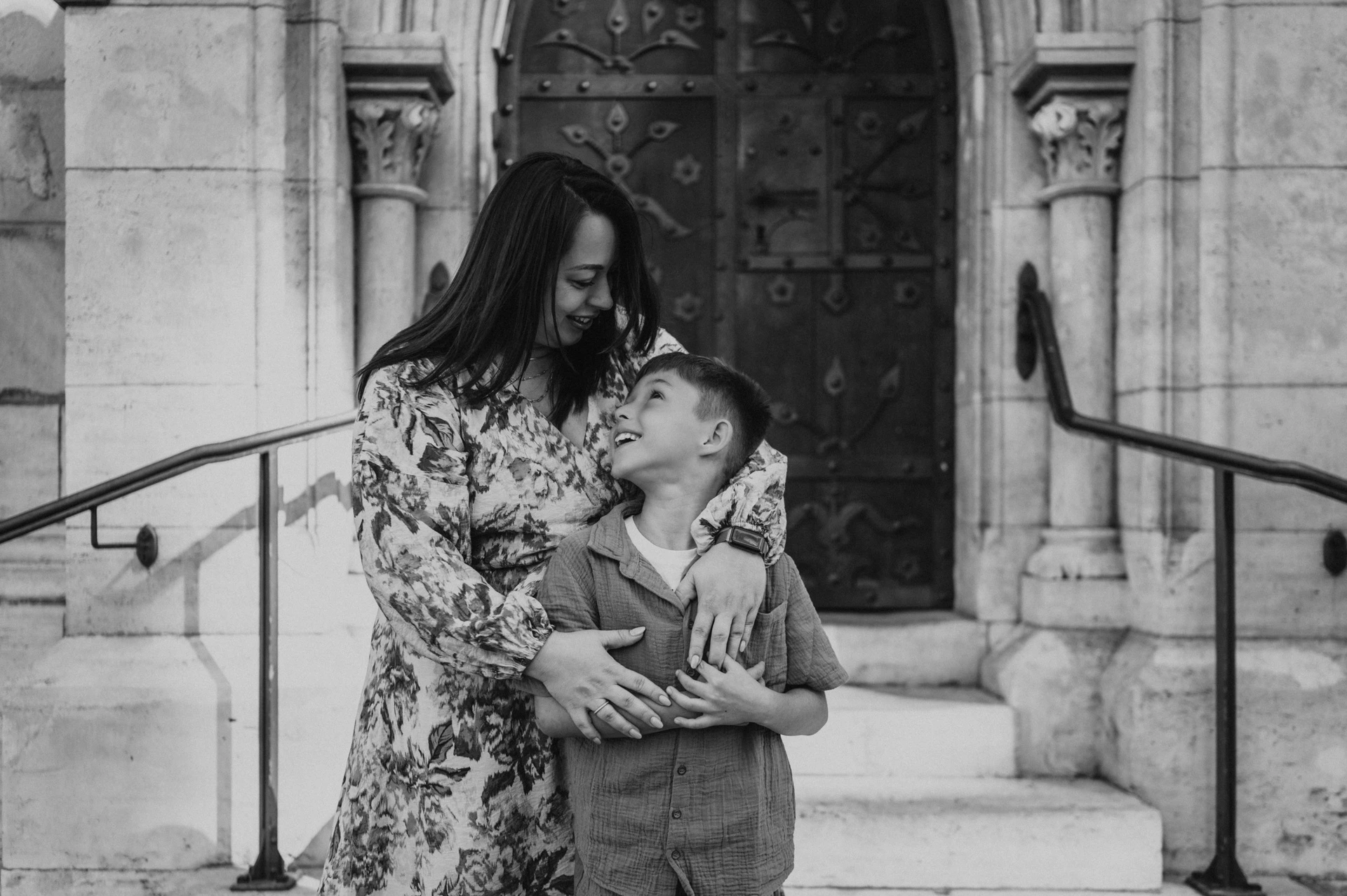 Smiles by the doorway in black and white | Budapest family photoshoot Black-and-white photograph of a mother embracing her son as he smiles up at her, with a carved wooden door and pale stone steps behind them during their family photoshoot in Budapest.