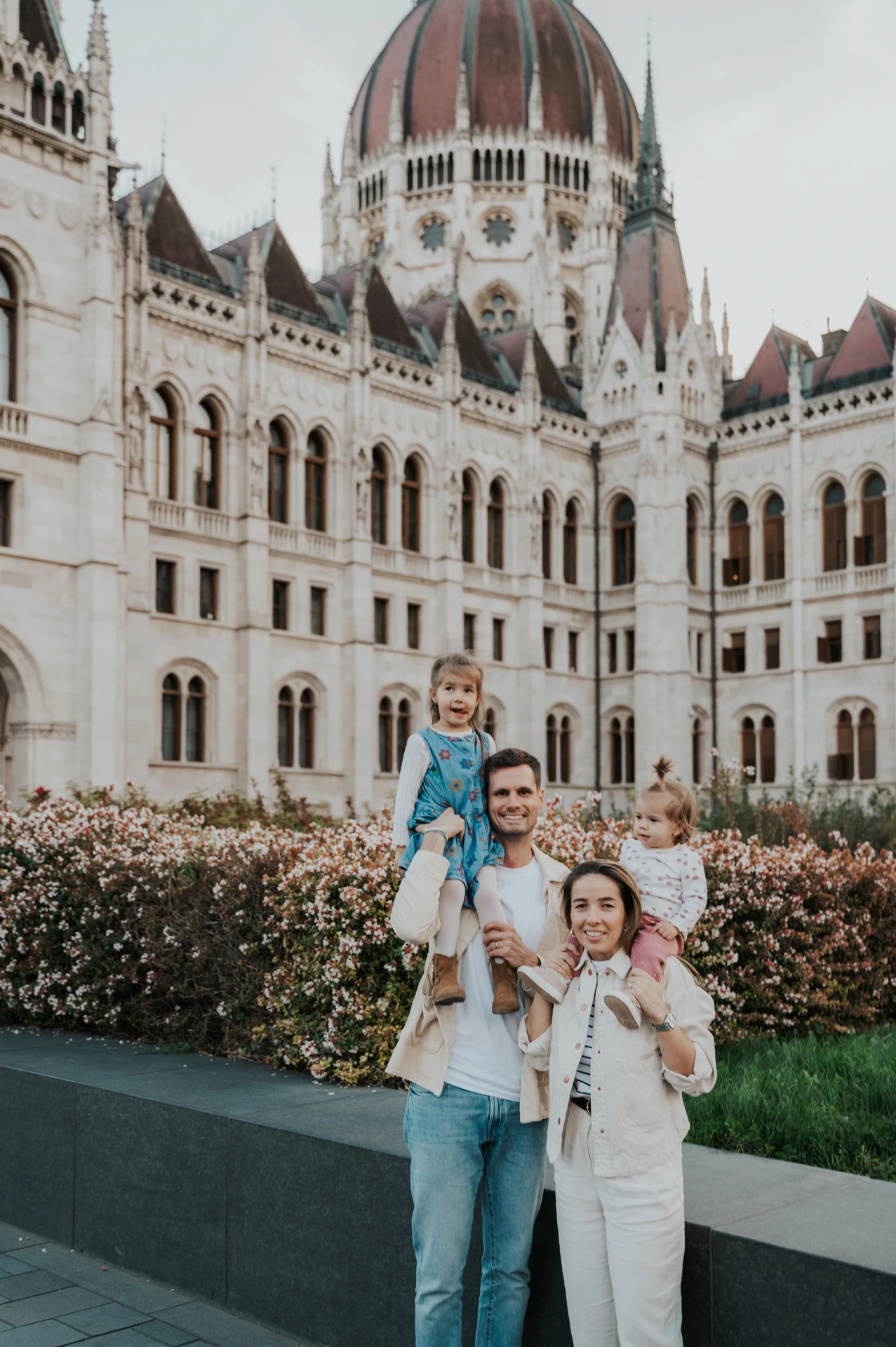 The parents carrying their two children beside the flowerbed during their Budapest family photoshoot, with Parliament’s dome rising above them.