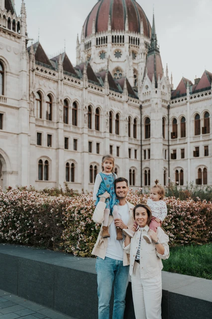 Budapest family photoshoot carrying the children by Parliament The parents carrying their two children beside the flowerbed during their Budapest family photoshoot, with Parliament’s dome rising above them.