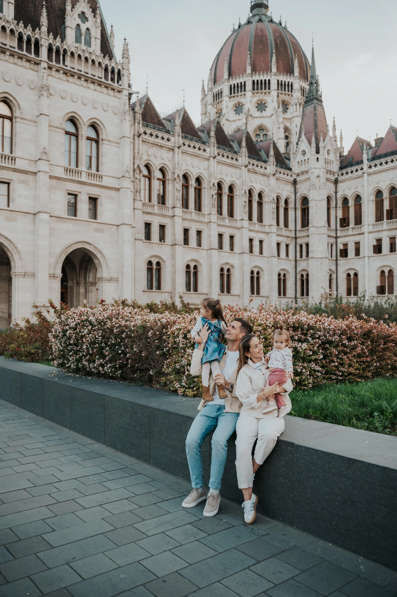 A parent lifting a child above the seated family during their Budapest family photoshoot, with Parliament’s dome and stonework behind them.