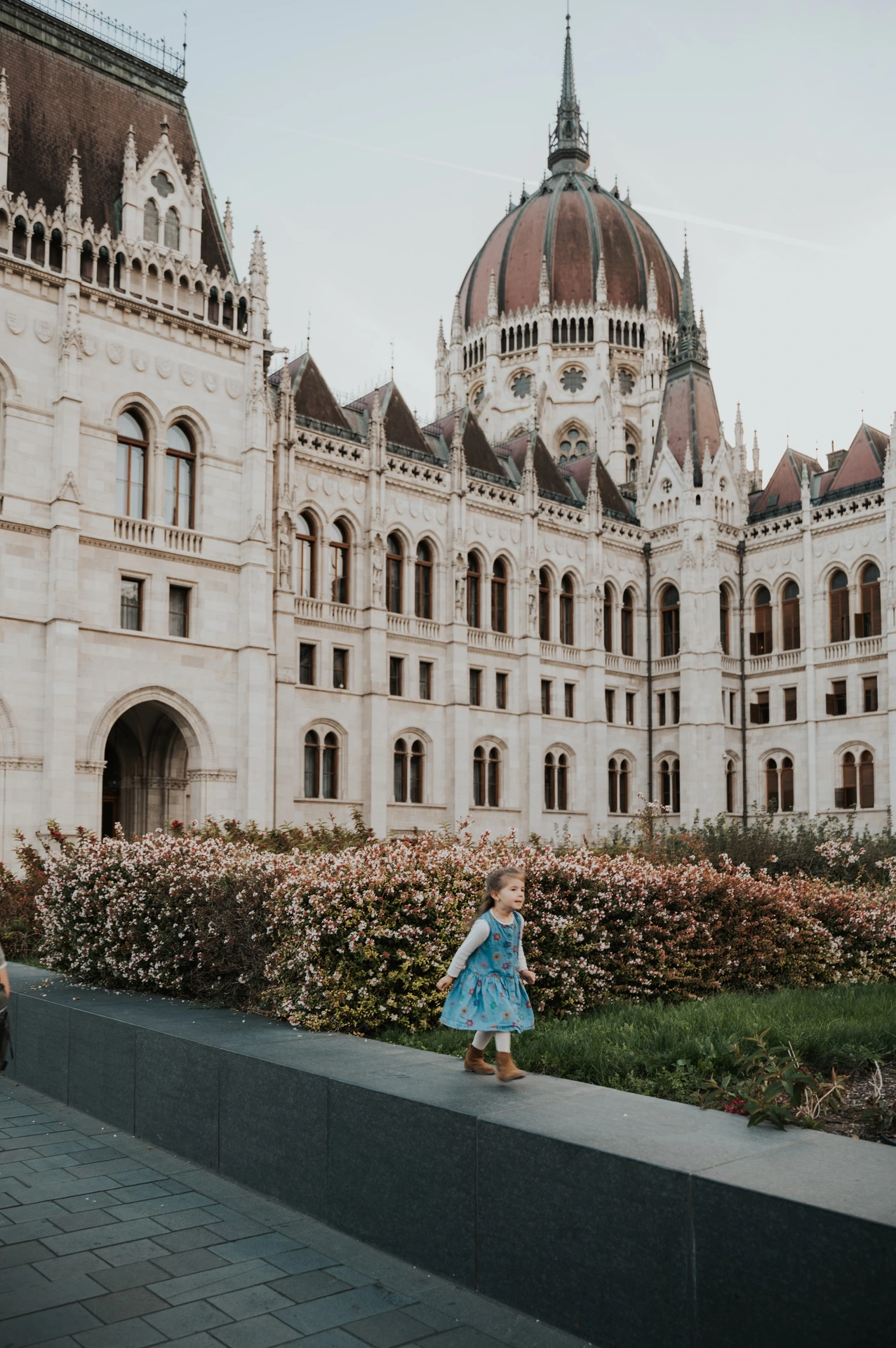 A child in a blue dress standing on a low stone ledge during the Budapest family photoshoot, with the Hungarian Parliament behind them.