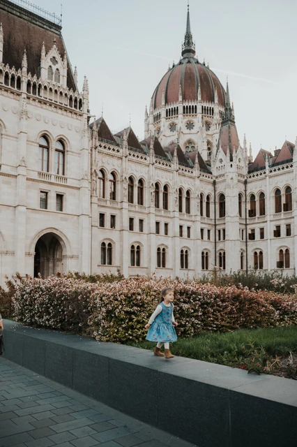 Budapest family photoshoot with a child on the Parliament ledge A child in a blue dress standing on a low stone ledge during the Budapest family photoshoot, with the Hungarian Parliament behind them.