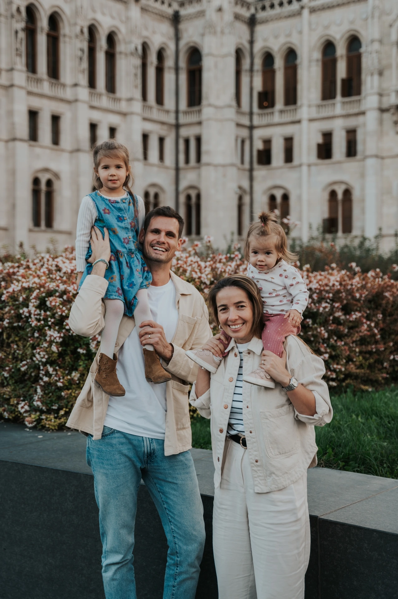 A close portrait of the parents smiling with their children on their shoulders during their Budapest family photoshoot, with Parliament behind them.