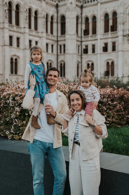Close Budapest family photoshoot with Parliament in the backdrop A close portrait of the parents smiling with their children on their shoulders during their Budapest family photoshoot, with Parliament behind them.