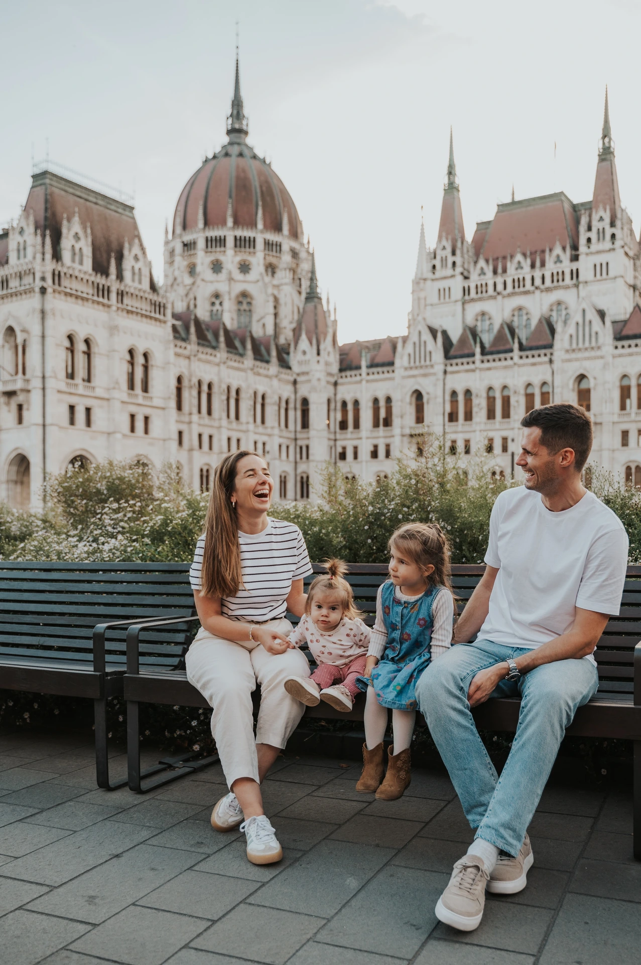 Budapest family photoshoot together by the Parliament bench The family gathered around a wooden bench during their Budapest family photoshoot, with one child held on a lap and Parliament behind the garden.