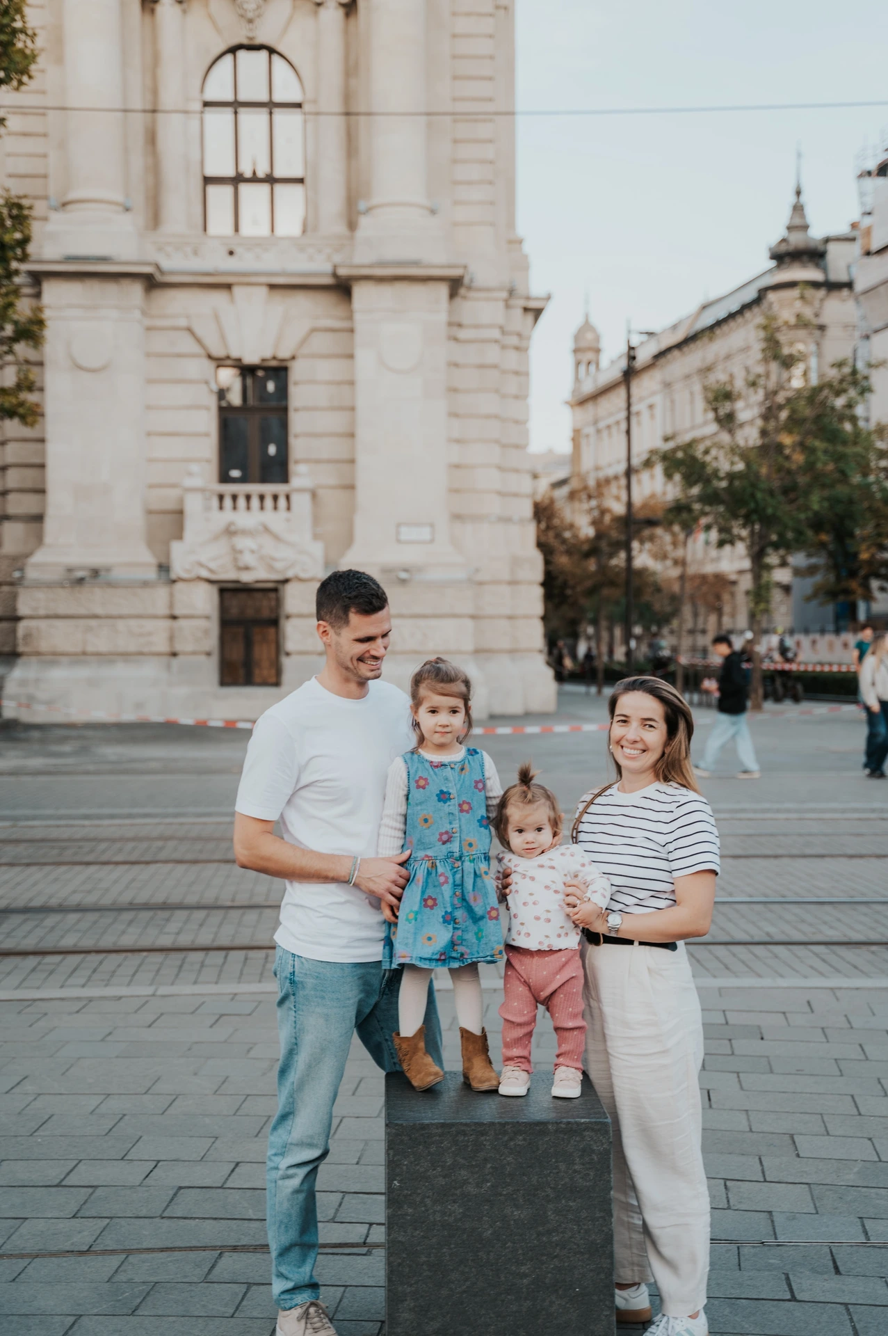 The parents looking towards their children during their Budapest family photoshoot beside a stone post, with historic city buildings in the background.