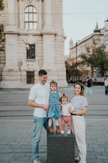 Budapest family photoshoot sharing a moment beside the stone post The parents looking towards their children during their Budapest family photoshoot beside a stone post, with historic city buildings in the background.