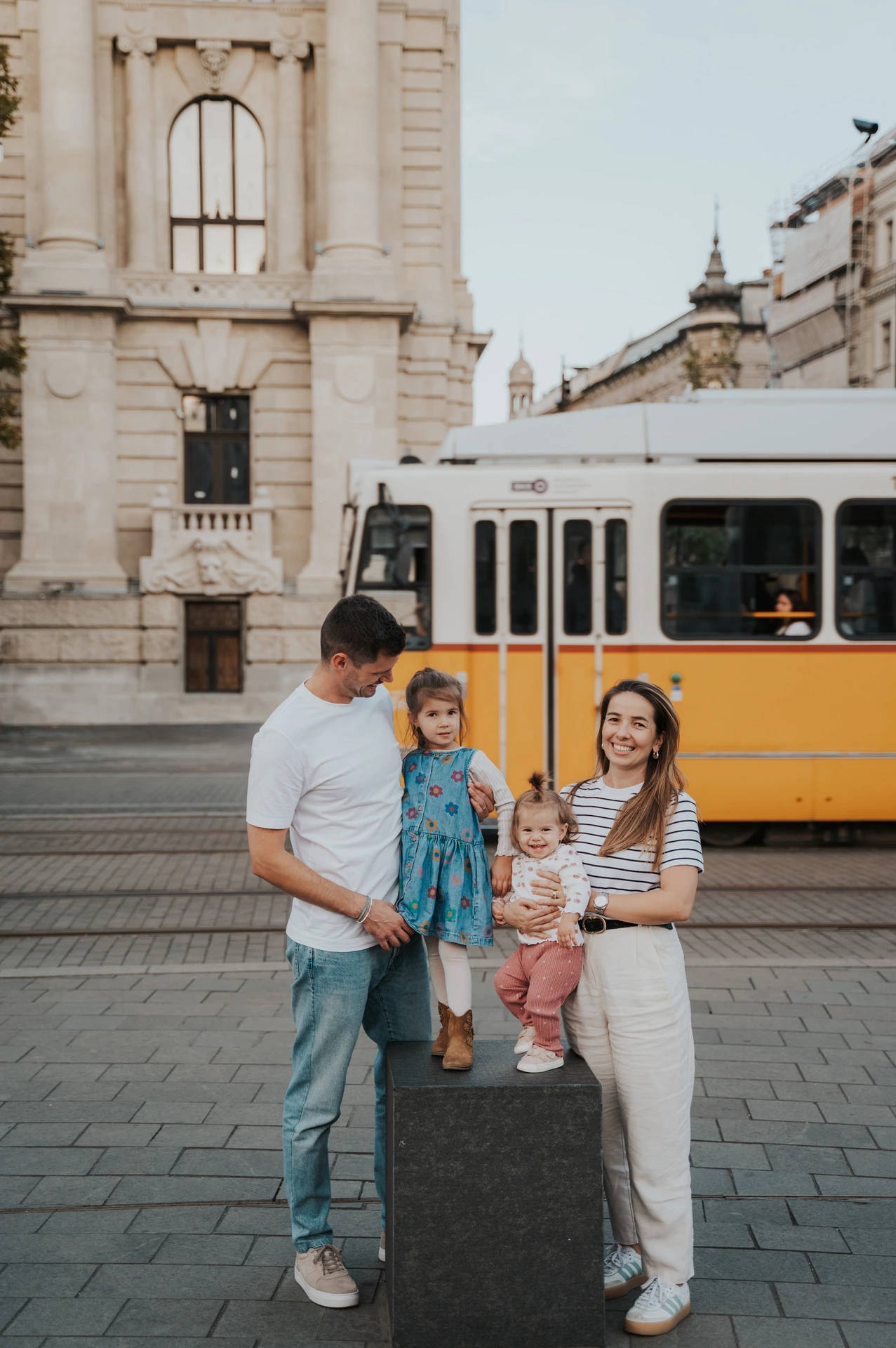 Budapest family photoshoot sharing smiles beside a passing tram The parents smiling towards their children during their Budapest family photoshoot beside the passing yellow tram, with both children on the stone post.