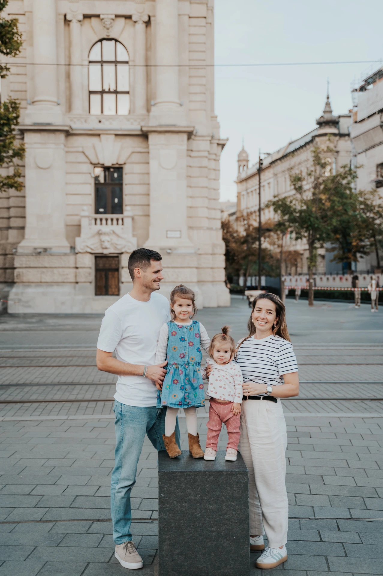 The parents standing beside their children on a low stone post during their Budapest family photoshoot, with city buildings behind them.
