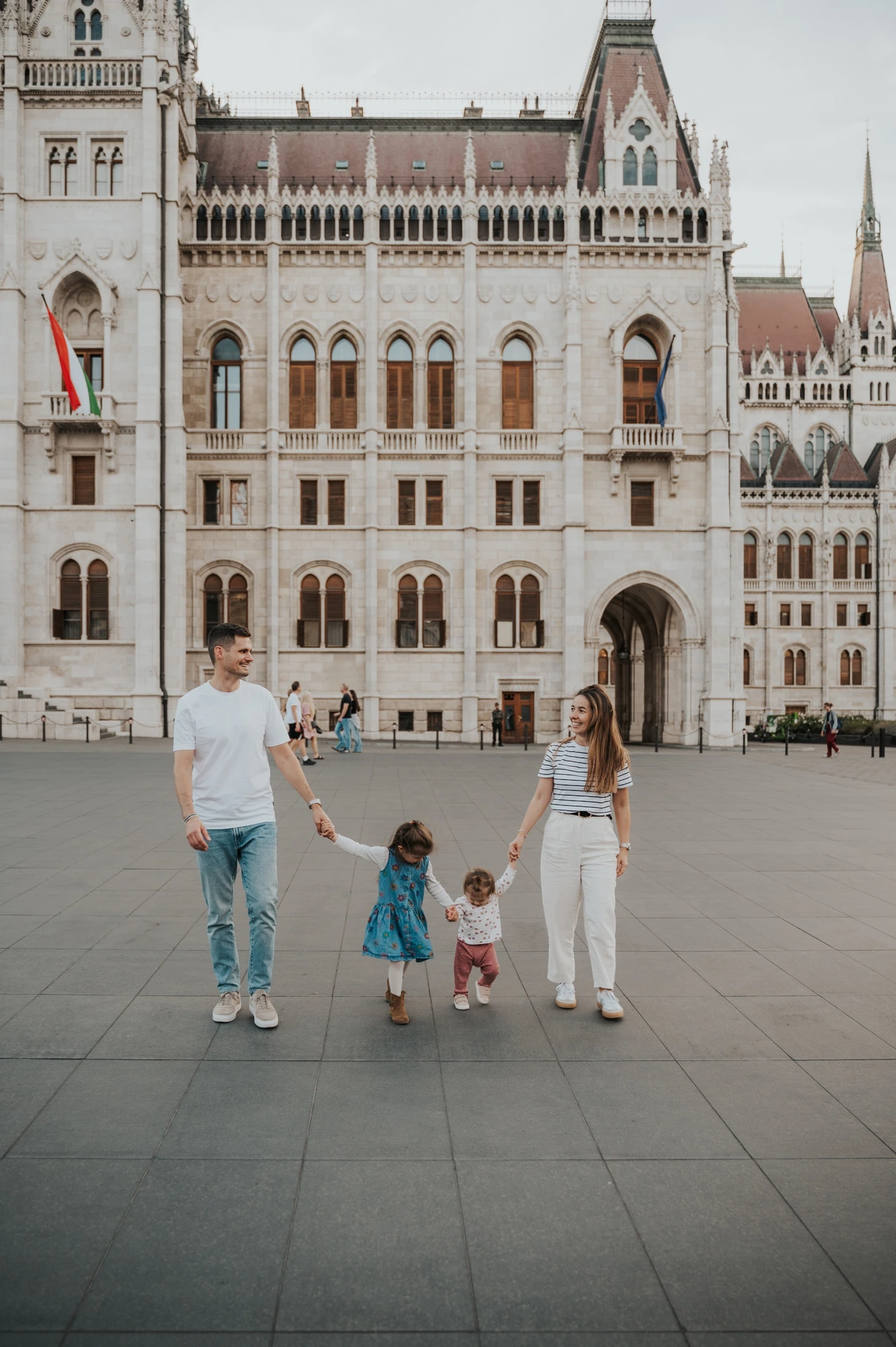 Family photoshoot in Budapest with a walk beside Parliament The parents and two children holding hands as they walk during their Budapest family photoshoot in front of Parliament’s tall stone façade.
