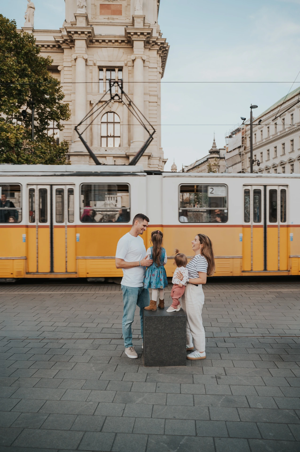 Budapest family photoshoot with a yellow tram in the backdrop The family together beside a stone post during their Budapest family photoshoot, with a yellow tram passing behind the parents and two children.