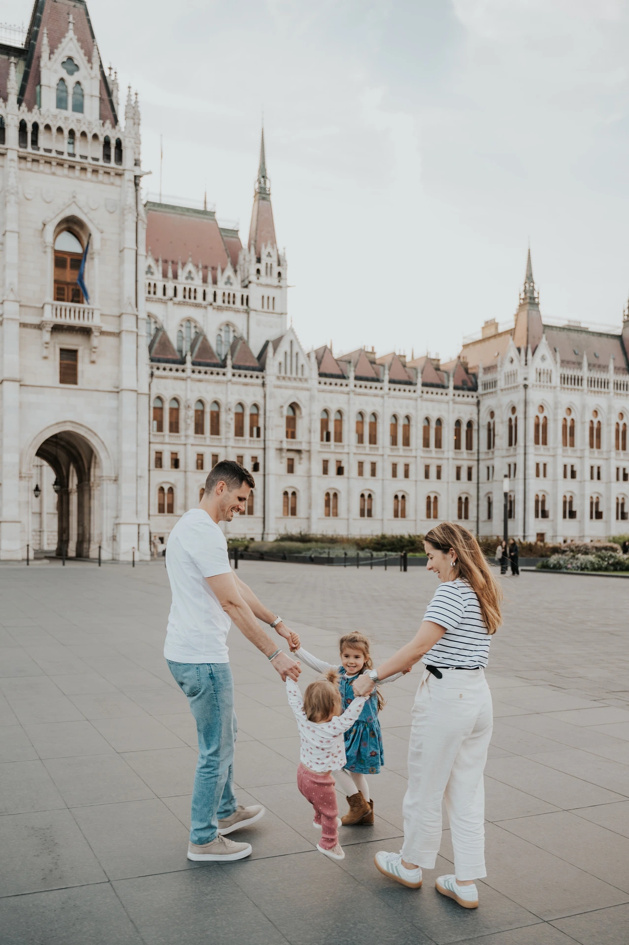 The parents holding hands with their two young children in the square during their Budapest family photoshoot, with Parliament’s façade behind them.