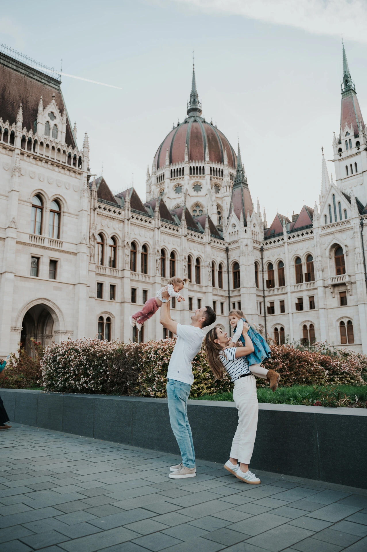 The parents lifting the children during a playful moment in their Budapest family photoshoot, with the Hungarian Parliament in the background.