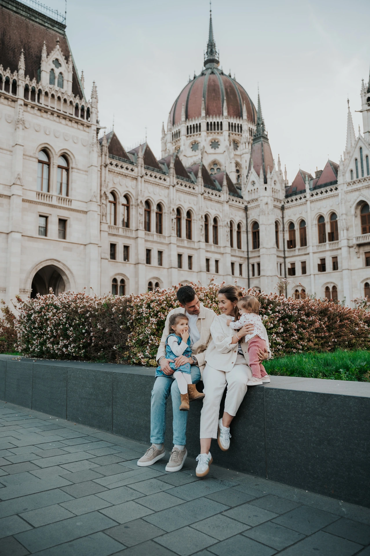The family sitting together on a low stone ledge during their Budapest family photoshoot, with the Hungarian Parliament and flowerbeds behind them.