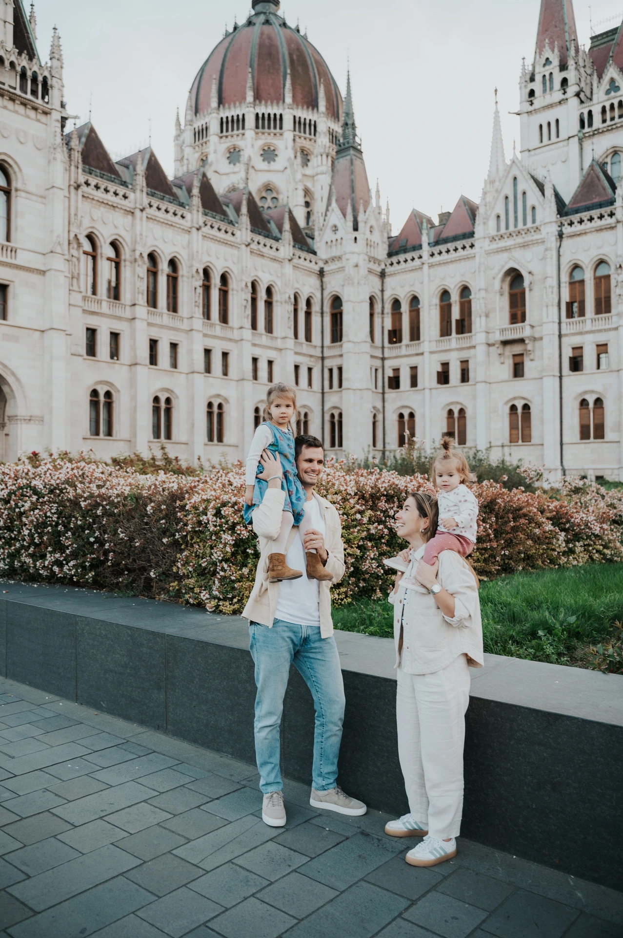 The parents looking towards each other while carrying the children on their shoulders during their Budapest family photoshoot at Parliament.