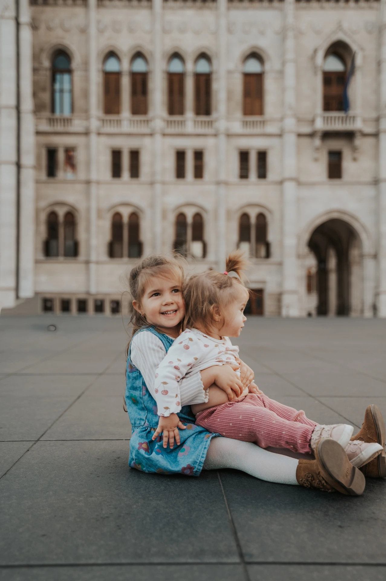 The two children sitting close together on the paving during their Budapest family photoshoot, one with an arm around the other and Parliament behind them.