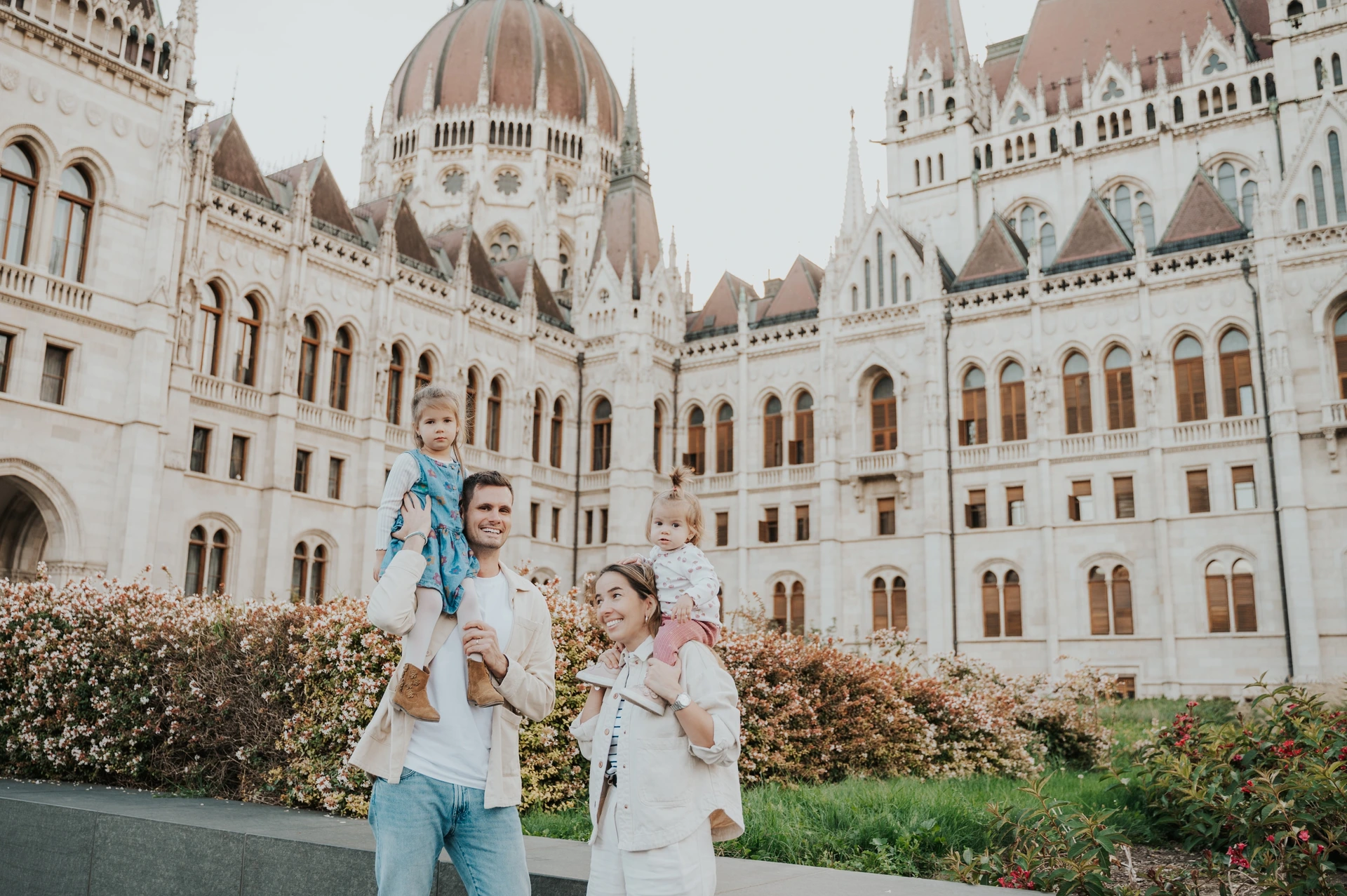 Budapest family photoshoot with Parliament’s dome and gardens Wide view of the family with both children on their parents’ shoulders during their Budapest family photoshoot, with Parliament’s dome and garden around them.