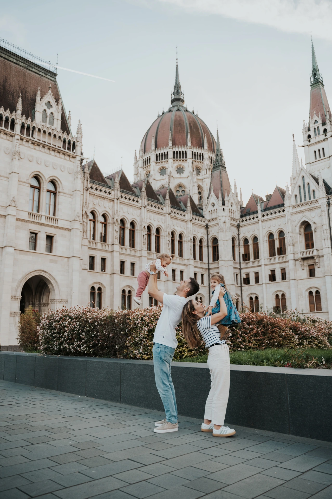 A wide view of the parents raising their children during their Budapest family photoshoot, with Parliament’s dome above the flowerbeds.
