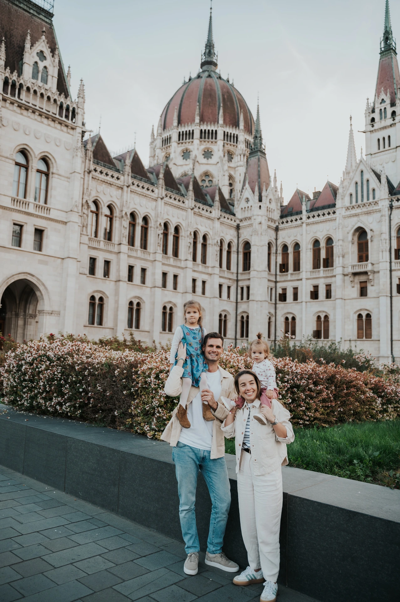 Budapest family photoshoot beneath Parliament’s dome Wide vertical view of the family during their Budapest family photoshoot, with the children on shoulders and Parliament’s dome in the backdrop.