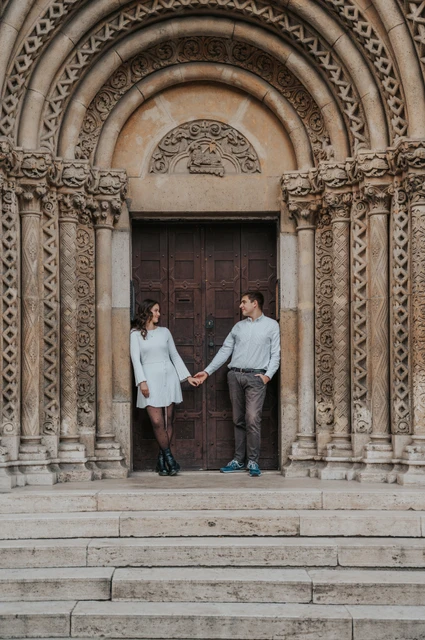 Holding hands beneath the castle arches at a Budapest anniversary photoshoot The couple holding hands beneath the carved stone arches at Vajdahunyad Castle during their anniversary photoshoot in Budapest.