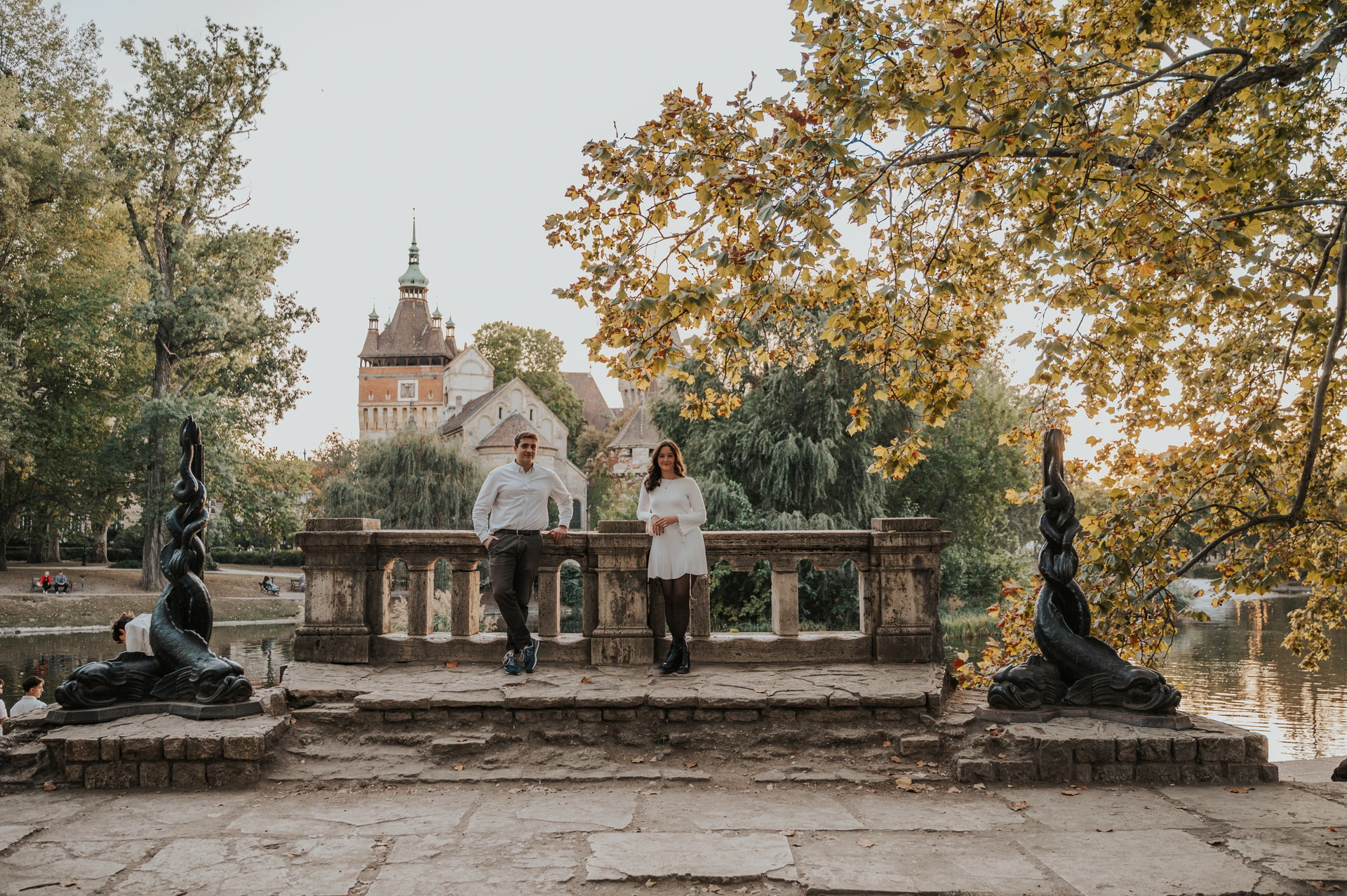 A wide view of the couple standing beside a stone balustrade between statues with Vajdahunyad Castle and golden leaves behind them during their anniversary photoshoot in Budapest.