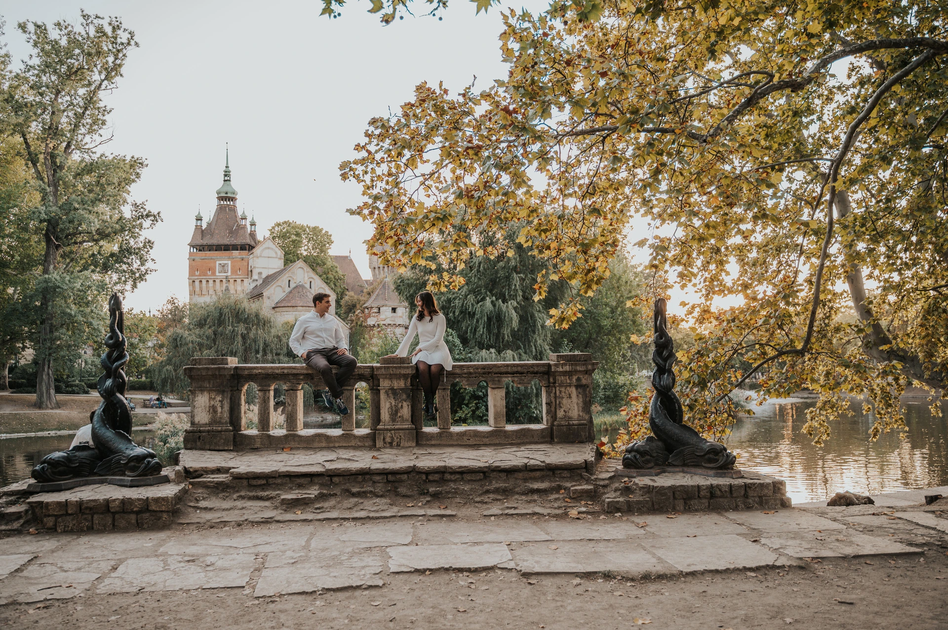 A castle view from the stone balustrade at a Budapest anniversary photoshoot A wide view of the couple sitting on a stone balustrade with Vajdahunyad Castle, statues and golden leaves around them during their anniversary photoshoot in Budapest.
