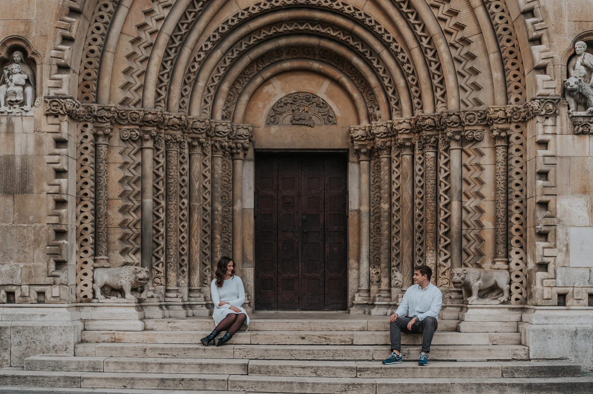 A landscape portrait of the couple sitting apart on the stone steps beneath Vajdahunyad Castle’s carved doorway during their anniversary photoshoot in Budapest.